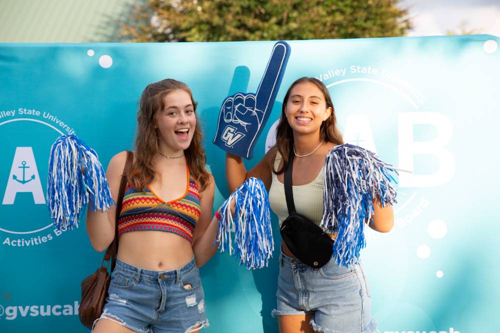 two students posing with foam fingers and pom poms in front of CAB backdrop at Laker Kickoff photo booth
